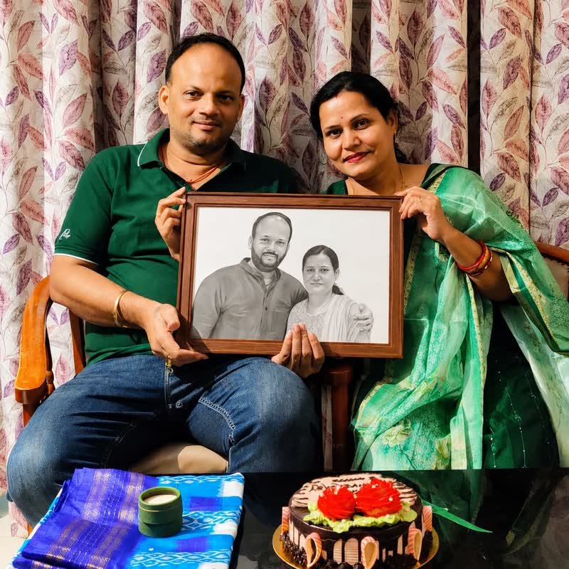 A couple holding a framed pencil portrait of the two of them, with a celebration cake on the table in front.