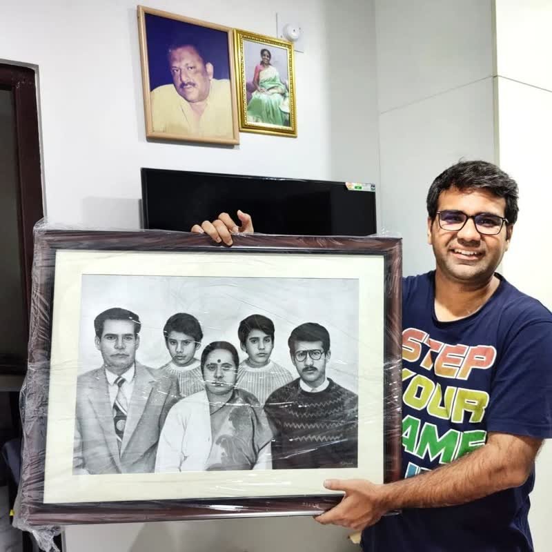 A customer holding a large framed pencil portrait of five family members, still wrapped in protective plastic, in his living room.