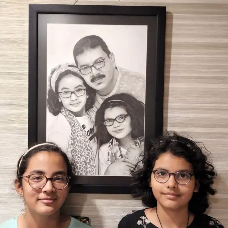 Two sisters standing beneath a framed pencil portrait of themselves with their father, hung on the wall of their home.